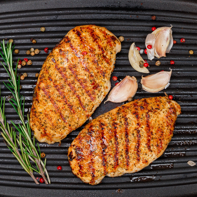 Grilled chicken breasts on a black grill pan with herbs and garlic cloves.